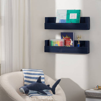 A cozy children's reading nook features a plush cream armchair with a blue and white striped blanket and a knitted shark toy. Two navy blue wall shelves hold books, markers, and a small trophy, while a model airplane and framed artwork add charm to the space.