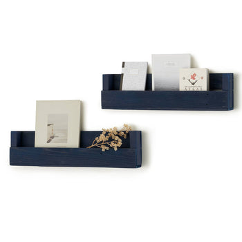 Two dark blue wooden wall-mounted shelves are displayed against a white background, holding books, magazines, and a small dried plant. The shelves have a rustic design with a front lip to keep the items in place.
