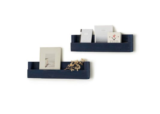 Two dark blue wooden wall-mounted shelves are displayed against a white background, holding books, magazines, and a small dried plant. The shelves have a rustic design with a front lip to keep the items in place.