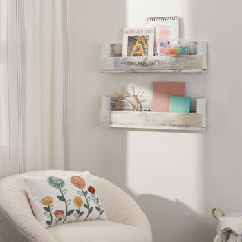 A cozy reading nook features a white boucle armchair with a floral embroidered pillow, positioned next to a wall with two rustic wooden shelves. The shelves hold books, notebooks, a small wooden decoration, and an art supply kit, adding a charming and functional touch to the space.
