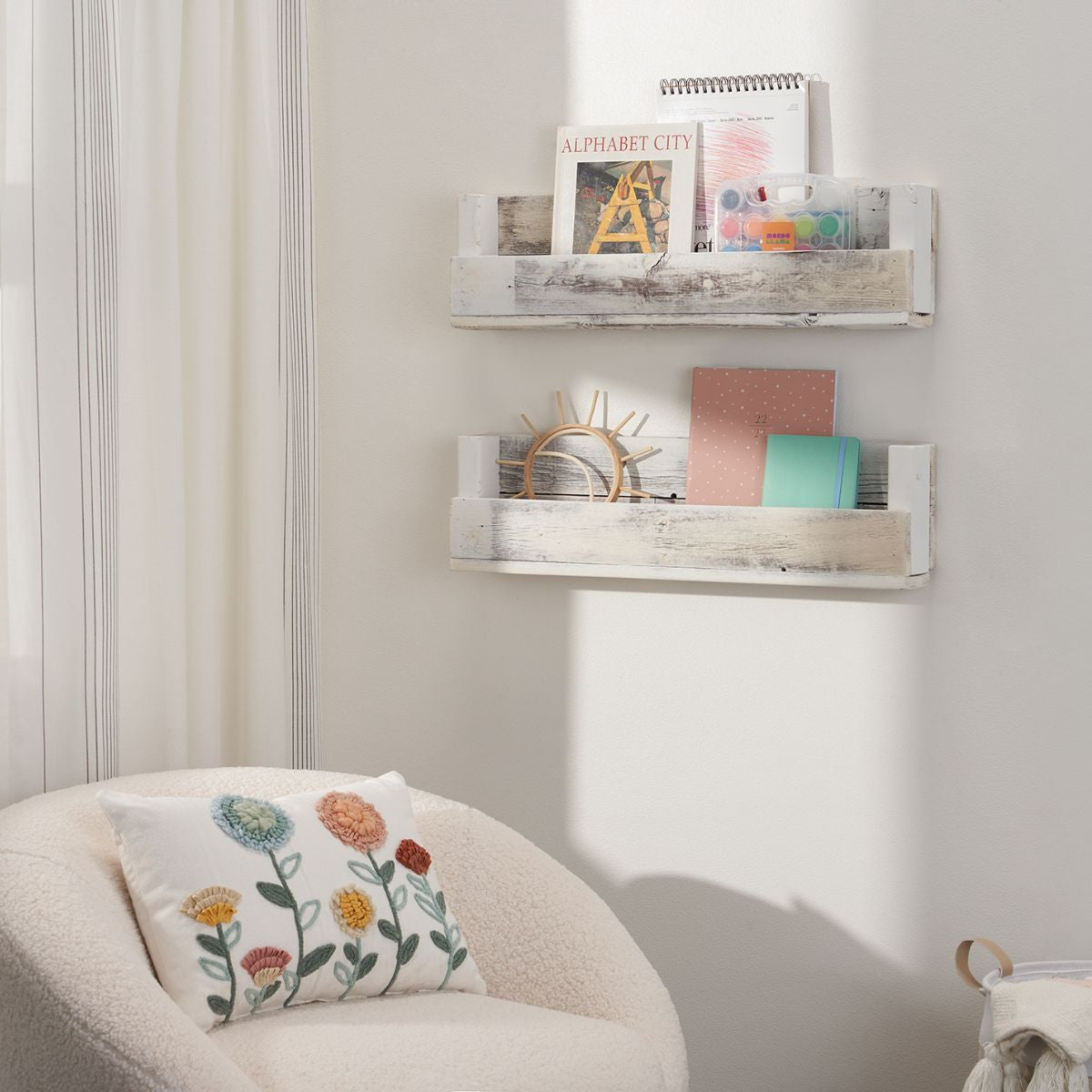 A cozy reading nook features a white boucle armchair with a floral embroidered pillow, positioned next to a wall with two rustic wooden shelves. The shelves hold books, notebooks, a small wooden decoration, and an art supply kit, adding a charming and functional touch to the space.