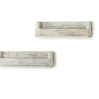 A set of two weathered white wooden shelves are mounted on a white wall in a minimalist composition. The shelves are rectangular with a small lip and appear to be floating, casting subtle shadows on the wall.