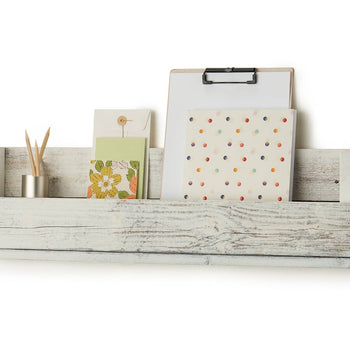 A white wooden shelf, made from reclaimed wood, is mounted on a white wall. The shelf has a rustic, weathered appearance and is holding a clipboard, a notebook, a greeting card, an envelope, and a small metal container with pencils.