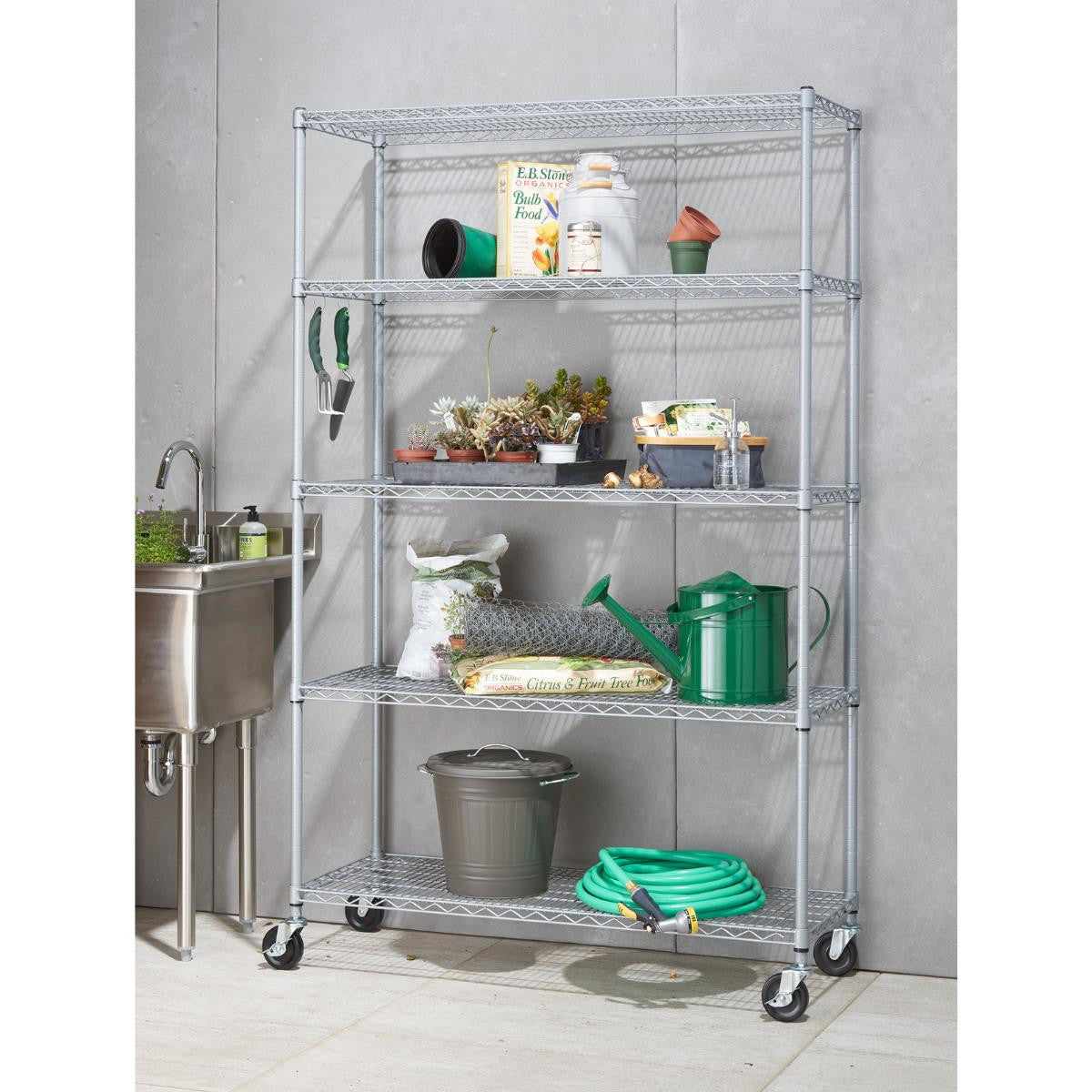 A gray wire shelving unit with five tiers stands next to a stainless steel utility sink. The shelves hold various gardening supplies, including potted plants, bags of soil, a watering can, a garden hose, and small tools.