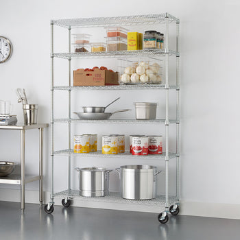 A six-tier chrome wire shelving rack in a commercial kitchen, filled with various kitchen supplies, stands against a white wall. The shelves hold items such as canned goods, bulk ingredients in clear containers, a box of tomatoes, onions in a wire basket, and large pots.