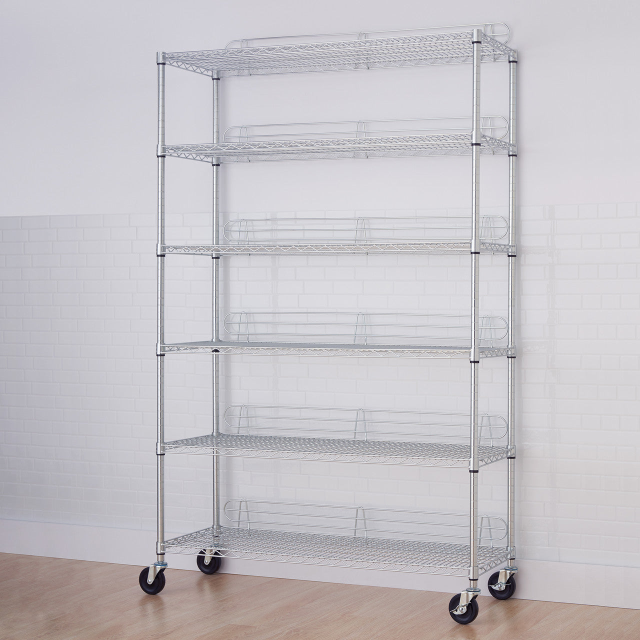 A 6-Tier Utility Shelving Unit with chrome shelving and wheels stands against a white brick wall. The wire shelves are visible, with some featuring a slight upward curve at the front, indicating a basket-like design.
