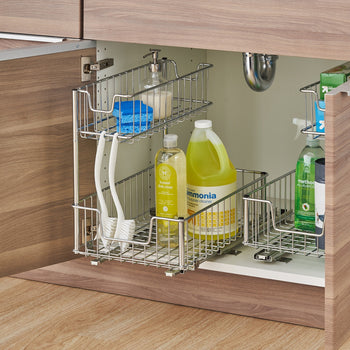 A chrome, two-tier pull-out organizer for an under-sink cabinet is filled with cleaning supplies. The top basket holds dish soap, a sponge, and a pump bottle, while the bottom basket contains larger bottles of dish soap and all-purpose ammonia, with two brushes hanging on the side.