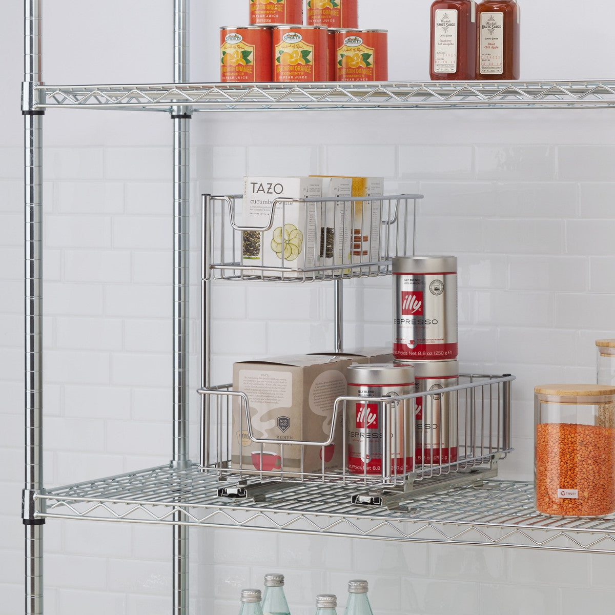 A chrome wire two-tier sliding undersink organizer is shown on a wire shelf. The top basket holds several boxes of Tazo tea, while the bottom basket contains a box and two red cans of Illy espresso.