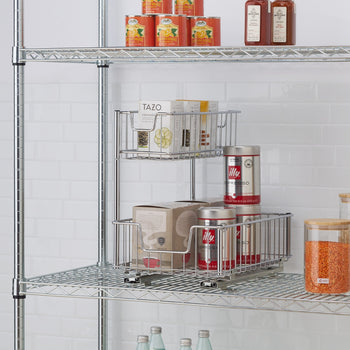 A chrome wire two-tier sliding undersink organizer is shown on a wire shelf. The top basket holds several boxes of Tazo tea, while the bottom basket contains a box and two red cans of Illy espresso.