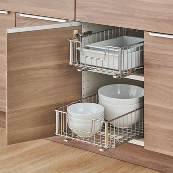 Two chrome wire pull-out drawers are installed in a wooden cabinet, each holding white dishes. The top drawer contains rectangular baking dishes, while the bottom drawer holds stacks of round bowls of various sizes.