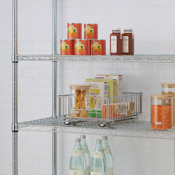 A chrome wire shelving unit holds various food items and kitchen organizers. On the middle shelf, a chrome wire pull-out drawer contains boxes of food and a clear canister of pasta.