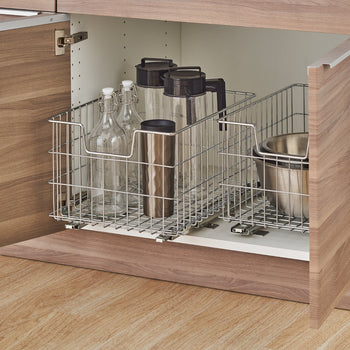 A chrome wire pull-out basket system is installed in a light wood cabinet, holding various kitchen items. The basket on the left contains glass bottles and a thermal mug, while the basket on the right holds a metal mixing bowl.