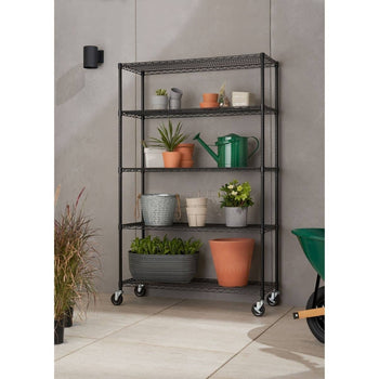 A black, five-tier wire shelf on wheels holds various potted plants and gardening supplies against a light gray wall. The shelving unit appears to be the Trinity 5-Tier Outdoor Wire Shelving, providing ample vertical storage for both indoor and outdoor use.