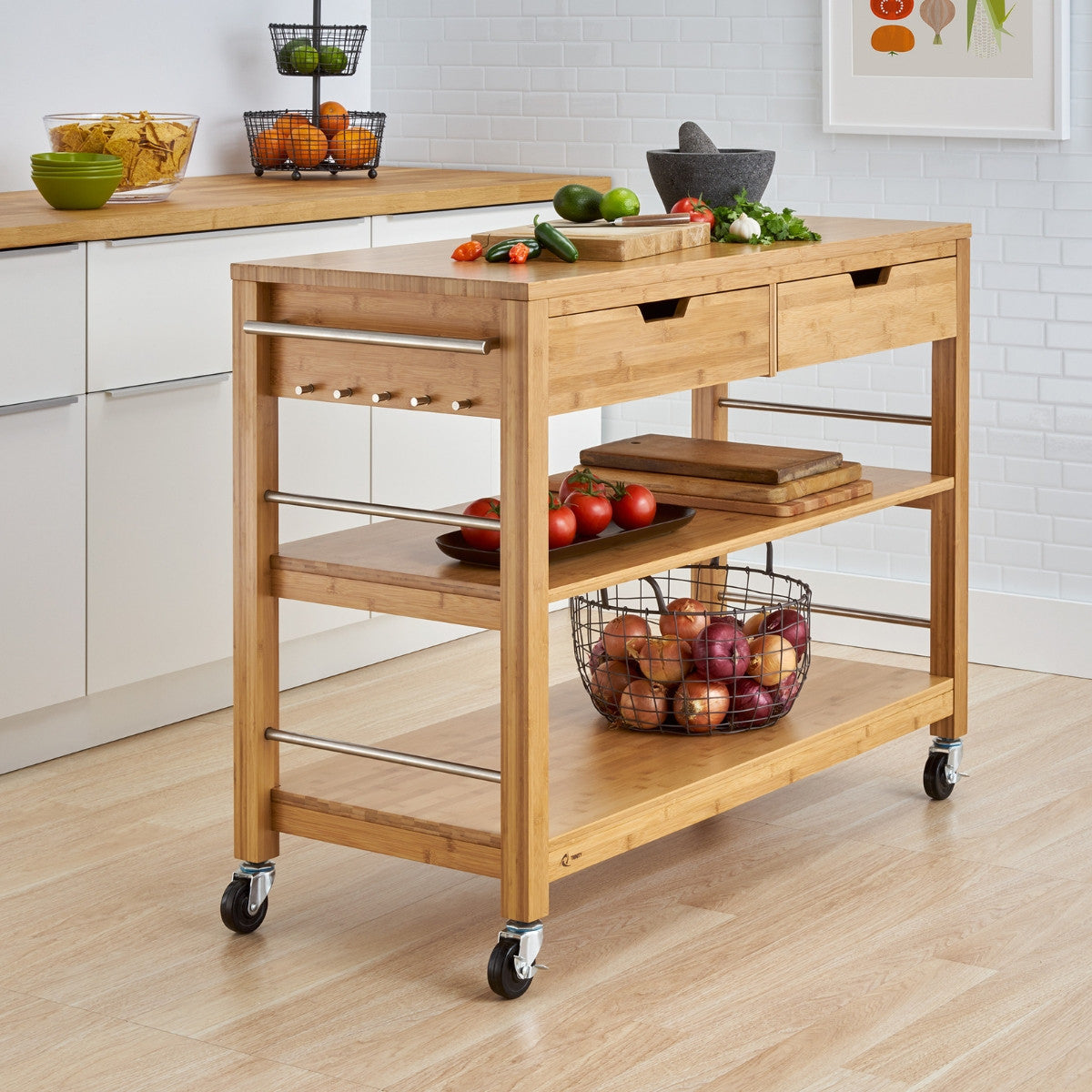 A natural wood kitchen island on wheels stands in a modern kitchen. The island features two drawers, two open shelves, a side rack with hooks, and a basket filled with onions.