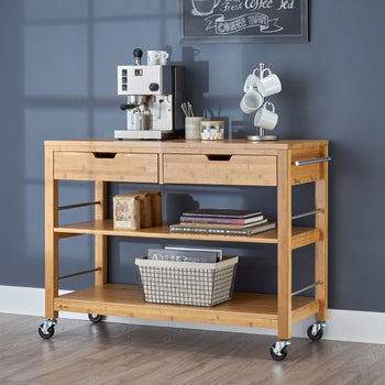 A natural wood kitchen island with two drawers and two open shelves on wheels. On top, there's an espresso machine, mugs, and a coffee accessories, while the shelves hold books, a basket, and bags of coffee.