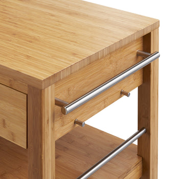 A close-up shot of the side of a bamboo kitchen island shows two stainless steel handles and two small bamboo pegs protruding from the side. The top surface and a lower shelf are visible, both made of light-colored bamboo wood.