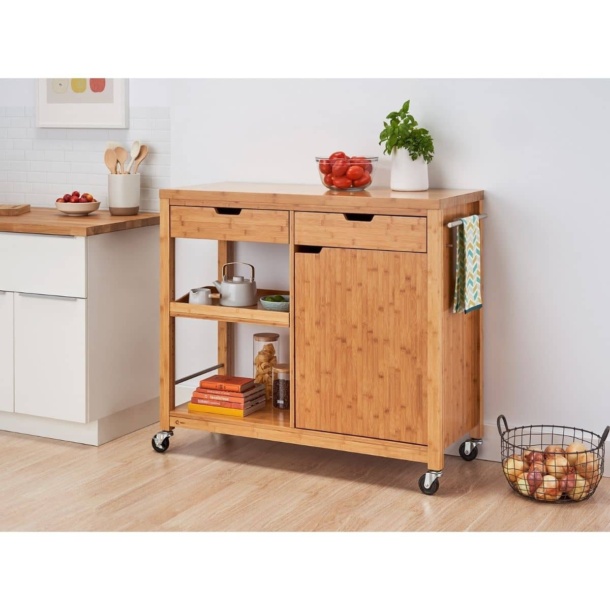 A bamboo kitchen island with drawers, a cabinet, and shelves stands on a light wood floor against a white wall. The cart holds various kitchen items, including tomatoes, a potted plant, cookbooks, and a metal teapot.
