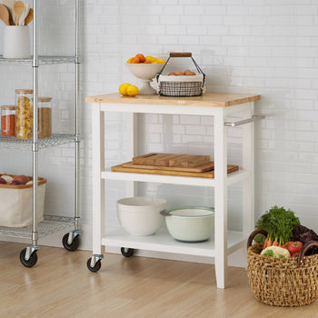 A white wooden kitchen cart with a light wood top stands in a kitchen with white subway tile. The cart has two open shelves, and a metal rack with wheels is visible to its left.