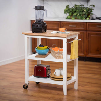 A white rolling kitchen cart with a wooden top holds a blender, bowls of fruit, a basket of oranges, a toaster, cups, and serving trays. A side-mounted towel bar with a yellow towel adds convenient storage and functionality.