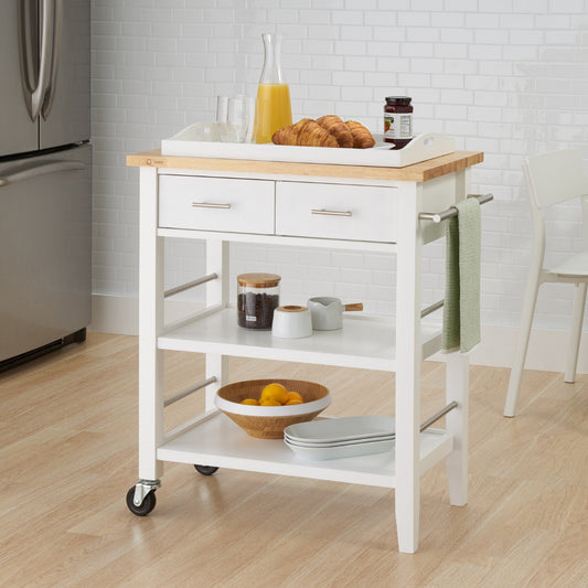 A white wooden kitchen cart with a light wood top stands on a light-colored wooden floor. The cart has two drawers, two open shelves, two wheels on one side, and a towel rack, with a breakfast tray on top and various kitchen items stored on the shelves.