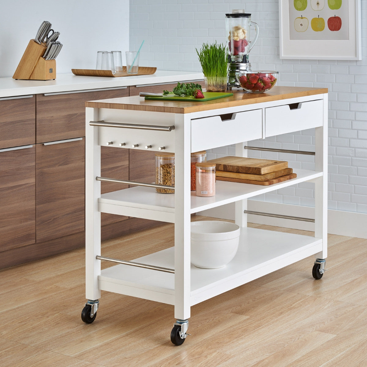 A white kitchen island with a light wood top stands on wheels, featuring two drawers, two open shelves, and a side with a towel bar and hooks. The top of the island is laden with fresh ingredients, a blender, and cutting boards, suggesting an active cooking space.