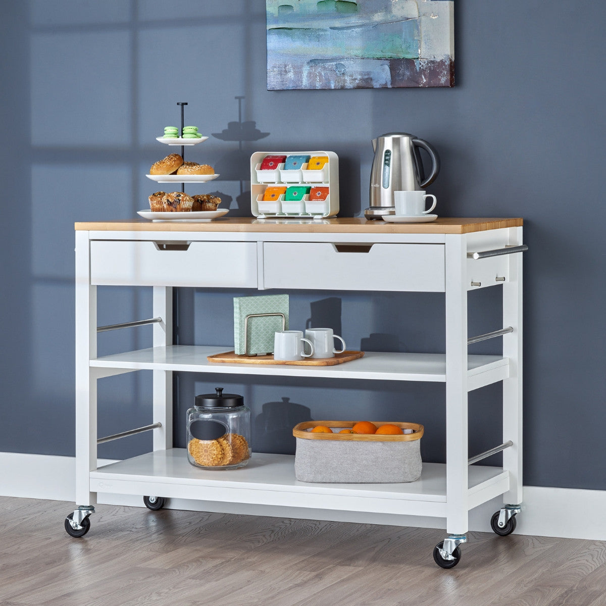A white kitchen island used as coffee bar. The rolling island cart features a light wood top, two drawers, two open shelves, and items like pastries, a kettle, mugs, and fruit.