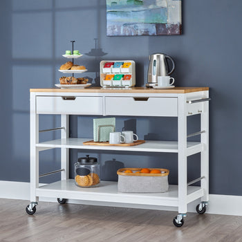 A white kitchen island used as coffee bar. The rolling island cart features a light wood top, two drawers, two open shelves, and items like pastries, a kettle, mugs, and fruit.