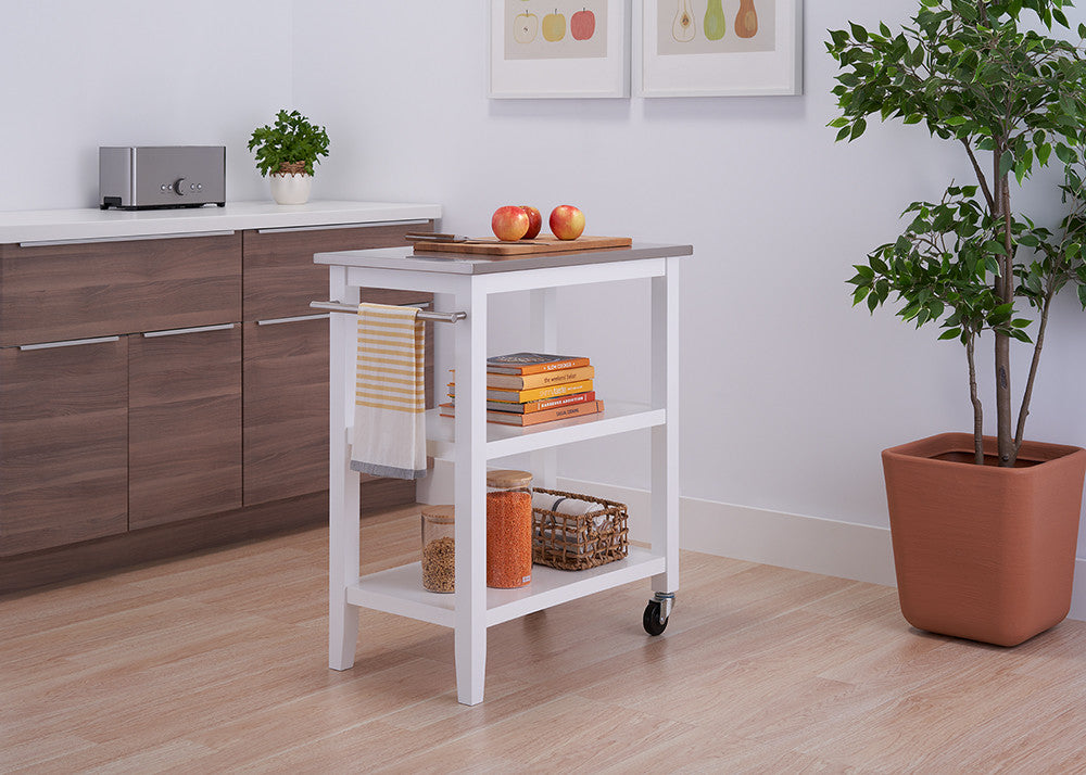 A white, three-tiered kitchen island cart with a stainless steel top is shown in a bright kitchen. The cart features two open shelves, a towel bar, and casters for mobility.