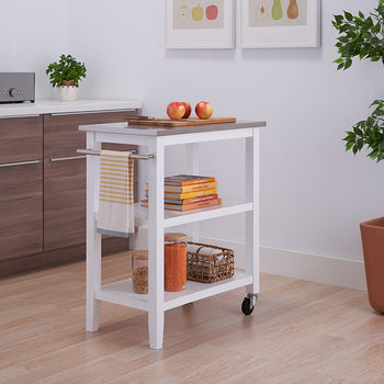 A white, three-tiered kitchen island cart with a stainless steel top is shown in a bright kitchen. The cart features two open shelves, a towel bar, and casters for mobility.