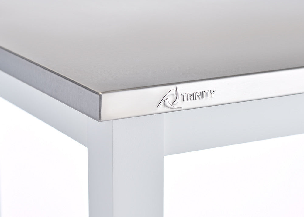 A close-up shot of a stainless steel tabletop with a beveled edge, featuring the "TRINITY" brand logo engraved on the surface. The table leg is white, and the background is blurred, focusing attention on the tabletop's material and branding.
