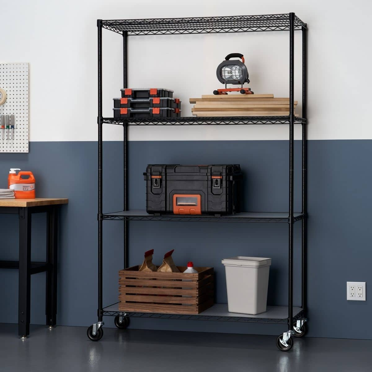 A black, four-tier wire shelving unit with wheels holds various items in a garage or workshop area. On the shelves are storage bins, tools, wood planks, a wooden crate, and a small trash can.