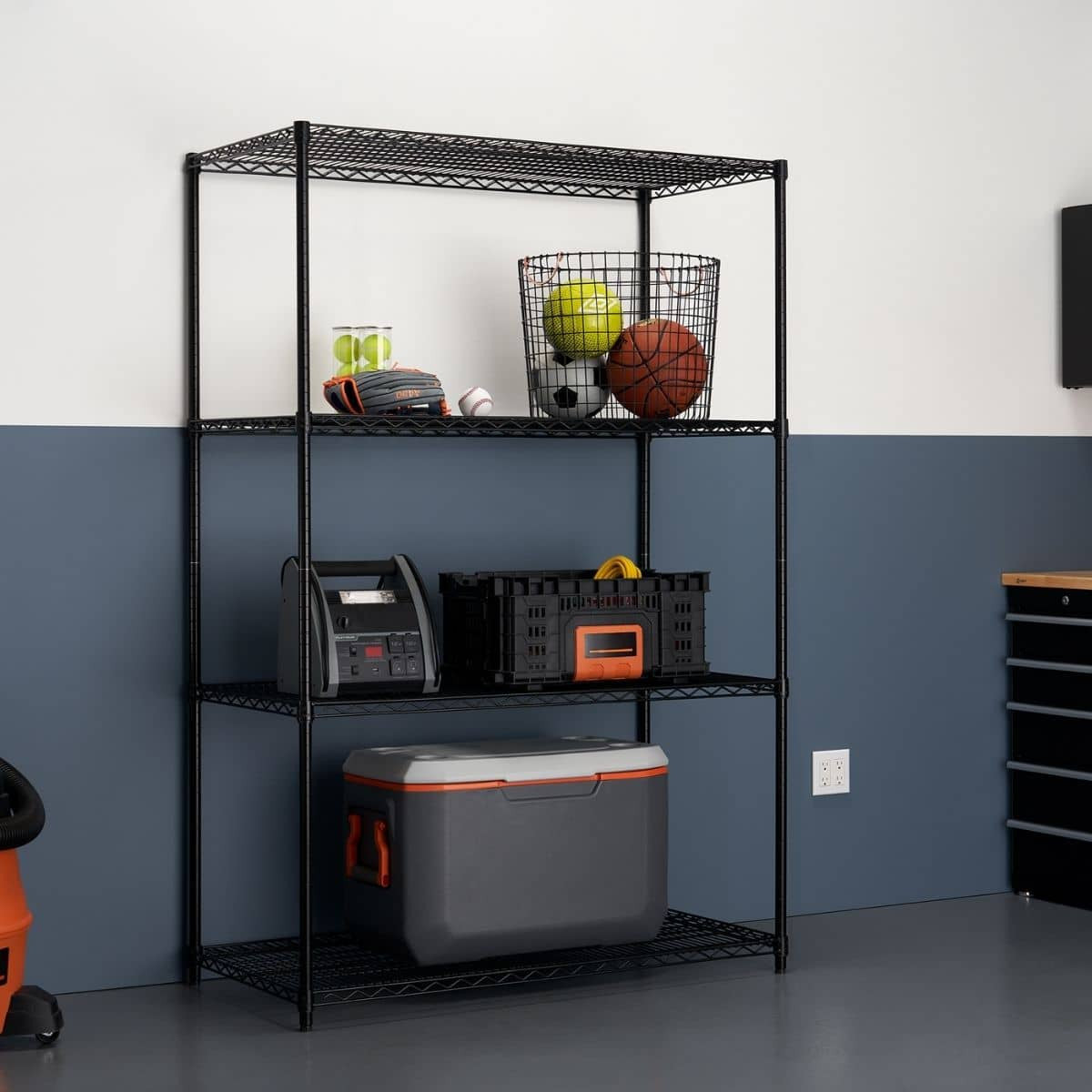 A black, four-tier wire shelving unit stands in a garage or storage area against a white and dark blue wall. The shelves hold various items including sports equipment in a basket, a battery charger, a black storage crate, and a gray and orange cooler.