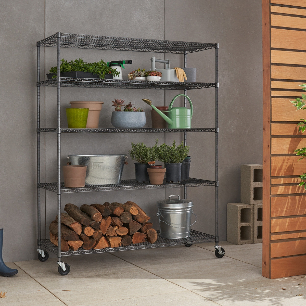 A tall, five-tier black wire shelving unit on wheels is shown against a light gray wall and a light-colored tiled floor. The shelves hold various gardening supplies, including potted plants, watering cans, tools, and bags of soil, along with a stack of firewood on the bottom shelf and a metal bucket.