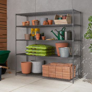 A metal shelving unit is stocked with various gardening supplies, including terracotta pots, watering cans, seed packets, bags of soil, and stacked bricks. The setup is organized neatly against a tiled wall, with a green wheelbarrow partially visible on the left side.