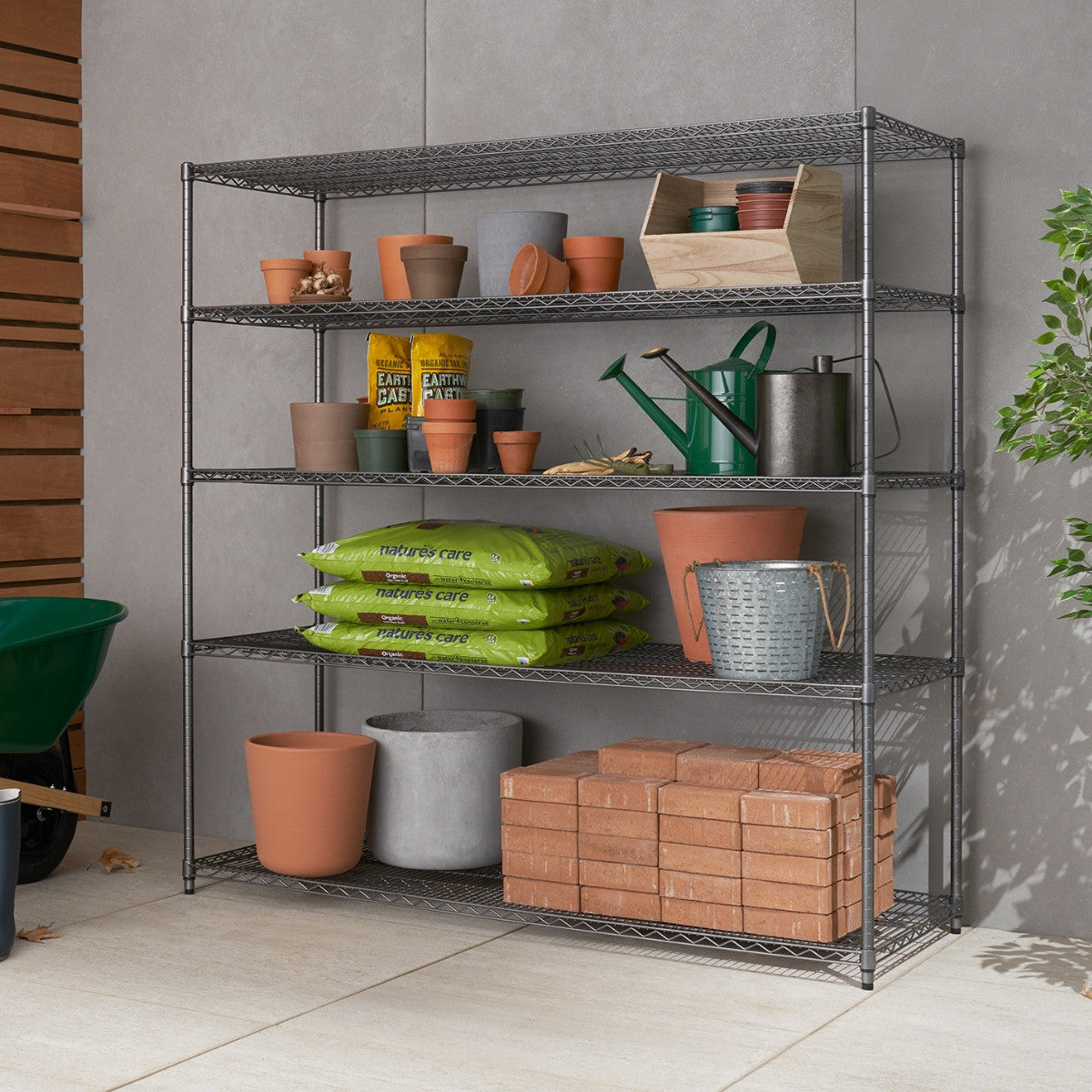 A metal shelving unit is stocked with various gardening supplies, including terracotta pots, watering cans, seed packets, bags of soil, and stacked bricks. The setup is organized neatly against a tiled wall, with a green wheelbarrow partially visible on the left side.