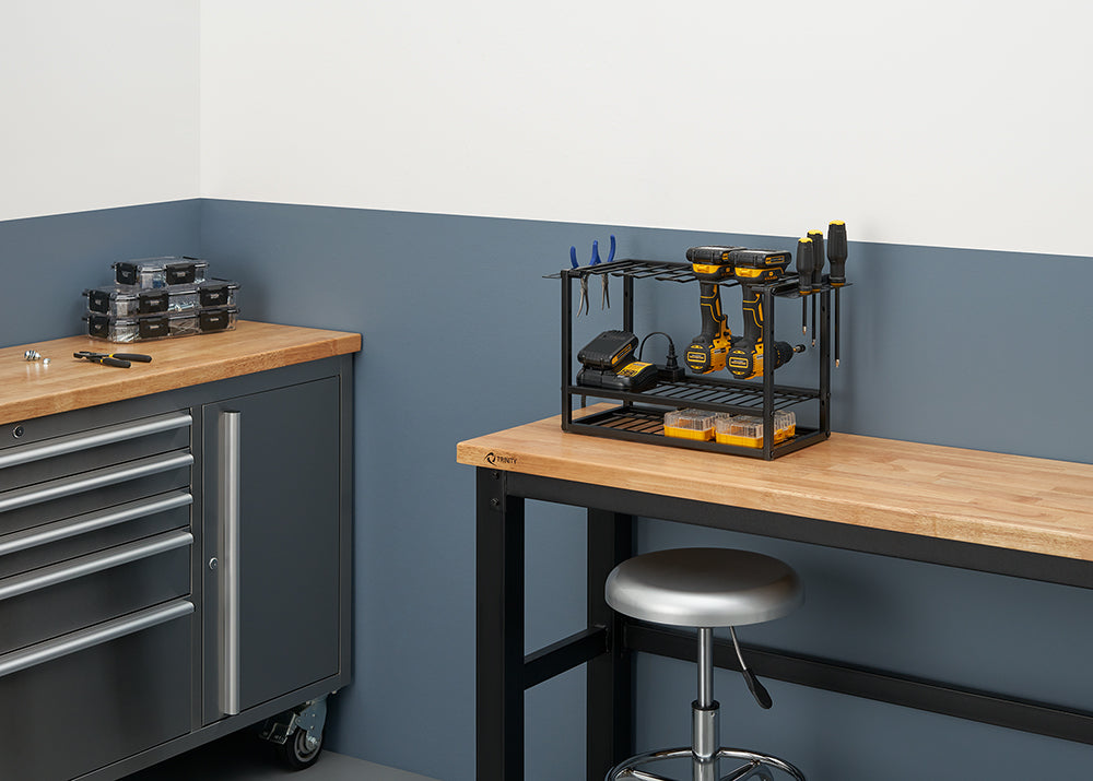 A workbench in a garage or workshop with a black metal organizer rack holding two cordless drills, batteries, and other tools. On the left, another workbench with drawers has several small parts storage boxes.
