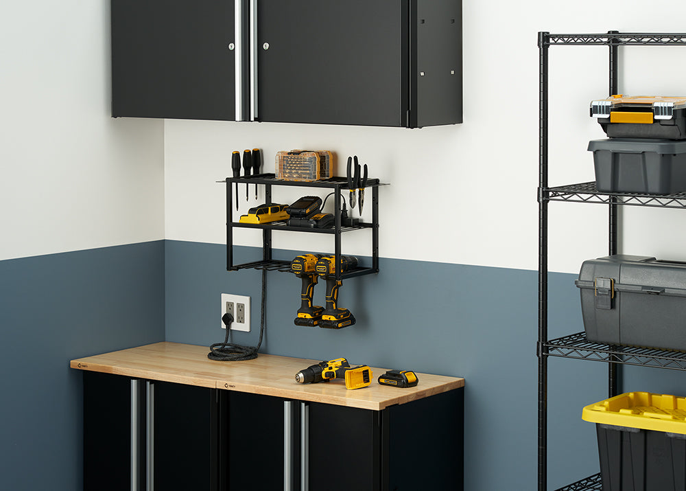 A well-organized garage workbench features a two-tone wall, with a light gray top half and a dark blue bottom half. A black metal shelf with hanging hooks holds two yellow and black cordless drills, two spare batteries, a charger, and various small hand tools.
