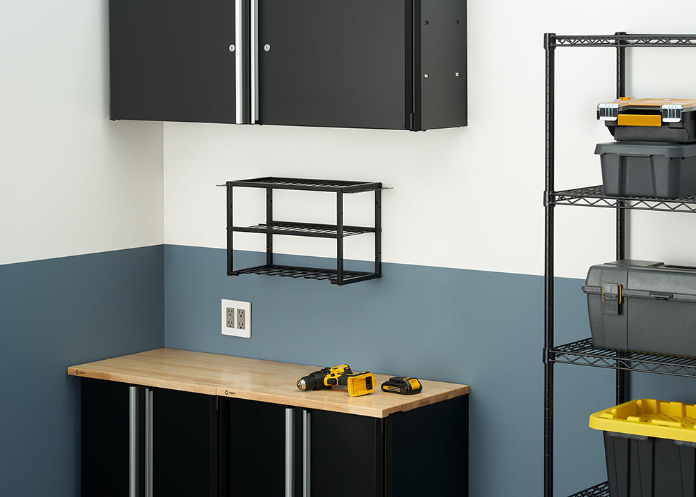 A neatly organized workshop or garage with a workbench and black cabinets against a two-toned wall. On the workbench, a drill and batteries are visible, while a black wire rack shelf is mounted on the wall above it.