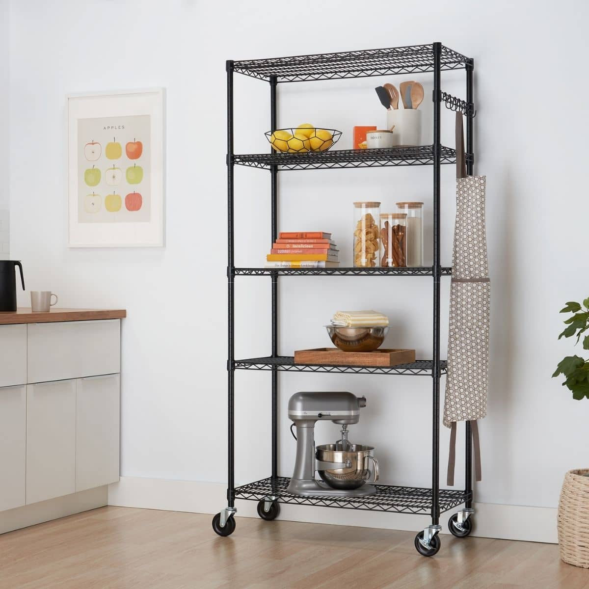 A black, five-tiered wire shelving unit on wheels stands against a white wall in a kitchen or pantry area. The shelves hold various kitchen items, including a stand mixer, bowls, jars, and fruit, and an apron hangs from the side.