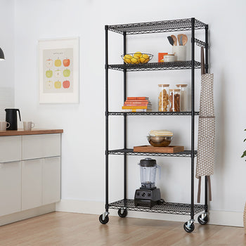 A 5-tier black wire shelving unit on caster wheels stands in a modern kitchen setting, organized with fruit, cookbooks, glass storage jars, utensils, and a blender. An apron hangs from side hooks on the rack, which is set next to white kitchen cabinets, a framed wall art piece, and a potted plant.