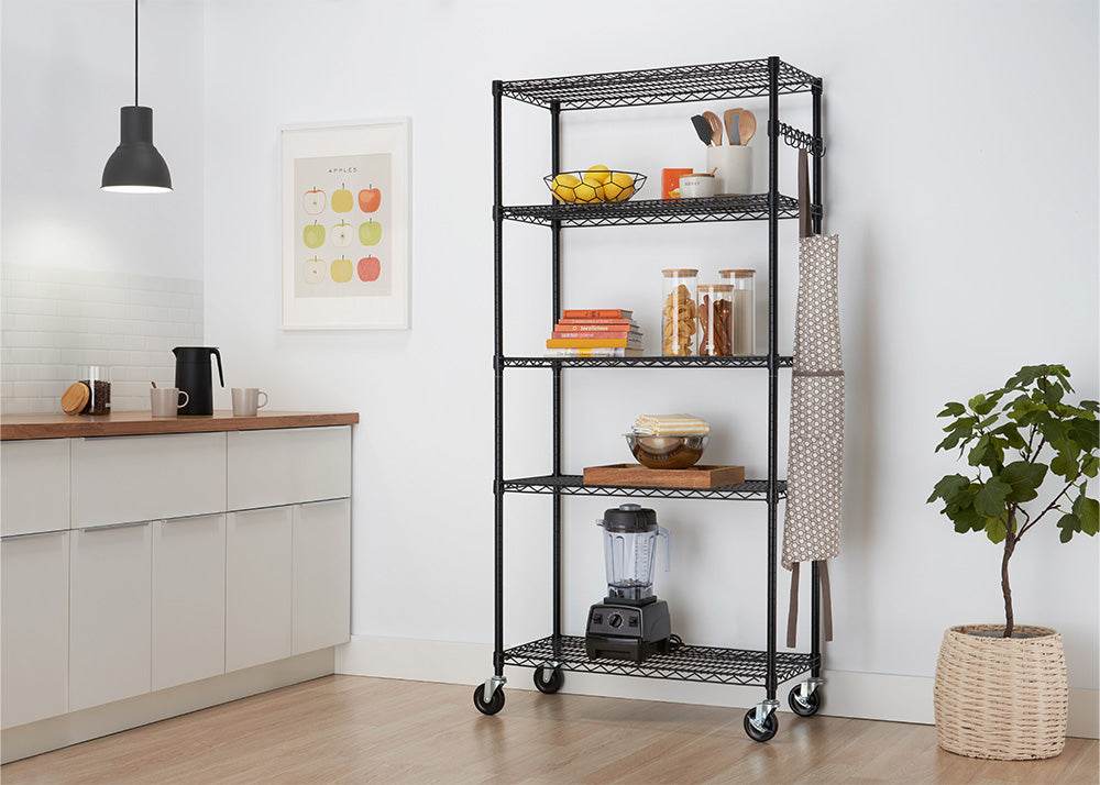 A 5-tier black wire shelving unit on caster wheels stands in a modern kitchen setting, organized with fruit, cookbooks, glass storage jars, utensils, and a blender. An apron hangs from side hooks on the rack, which is set next to white kitchen cabinets, a framed wall art piece, and a potted plant.