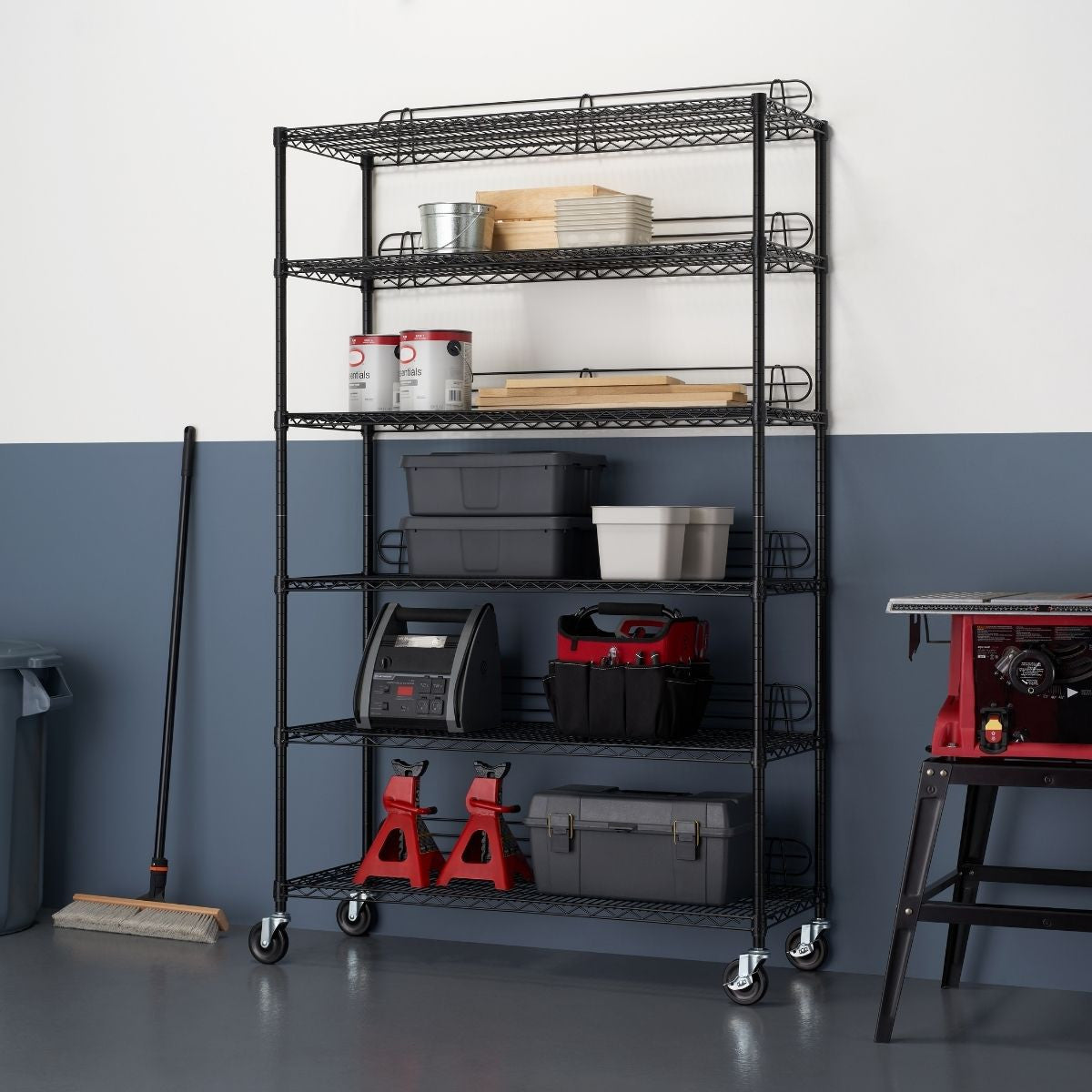 A tall, black wire shelving unit with six tiers stands against a two-toned wall, the lower half painted a deep blue and the upper half white. The shelves are filled with various storage containers, tools, and supplies, and the unit rests on wheels.