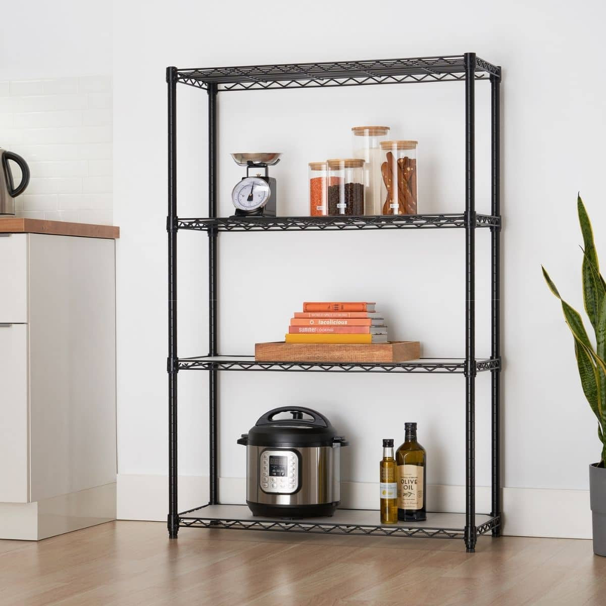 A black four-tier wire shelving unit stands against a white wall, holding various kitchen and household items. The shelving appears sturdy and functional, providing open storage in a room with a light wood floor.