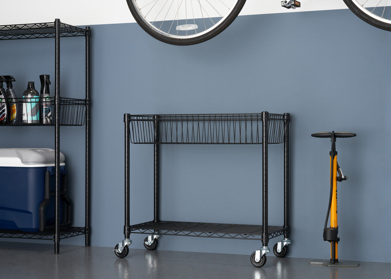 A black wire shelving unit with two tiers and wheels sits in the center of a garage against a dark blue wall, with a taller shelving unit to the left and a yellow bicycle pump to the right. A bicycle wheel hangs above.