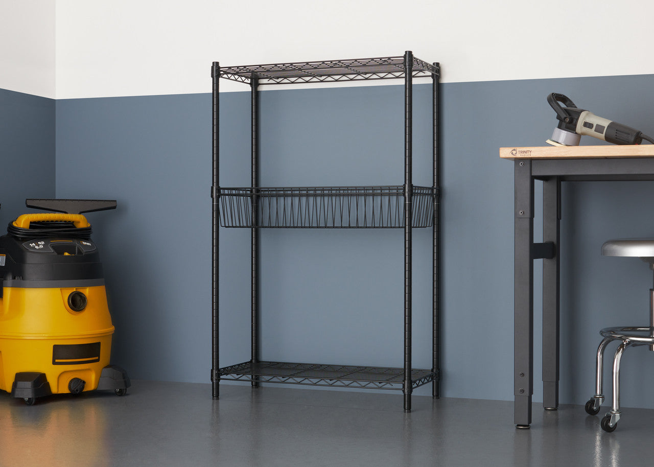 A black wire shelving unit with three tiers stands against a two-tone wall, featuring a basket on the middle shelf. To the left, a yellow wet/dry vacuum is visible, while a workbench and stool are on the right.