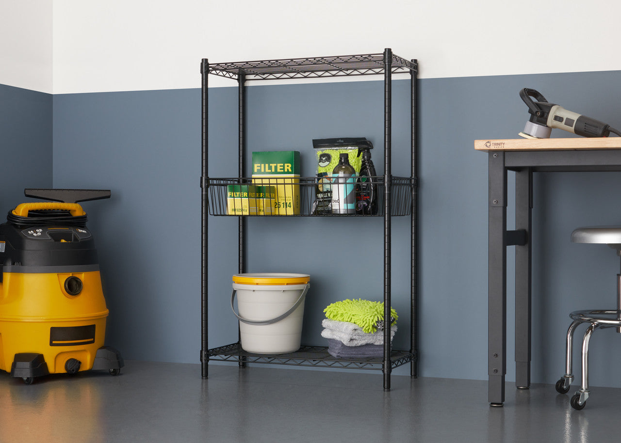 A black wire shelving unit with three tiers stands against a two-toned wall, holding various cleaning supplies. To the left, a yellow wet/dry vacuum is visible, and to the right, a metal work table with a stool is partially seen.