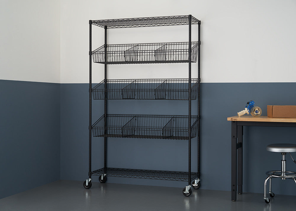 A black wire shelving unit with five tiers, some holding wire baskets, stands against a two-tone wall with a dark blue lower section. A wooden table and a metal stool are partially visible to the right.