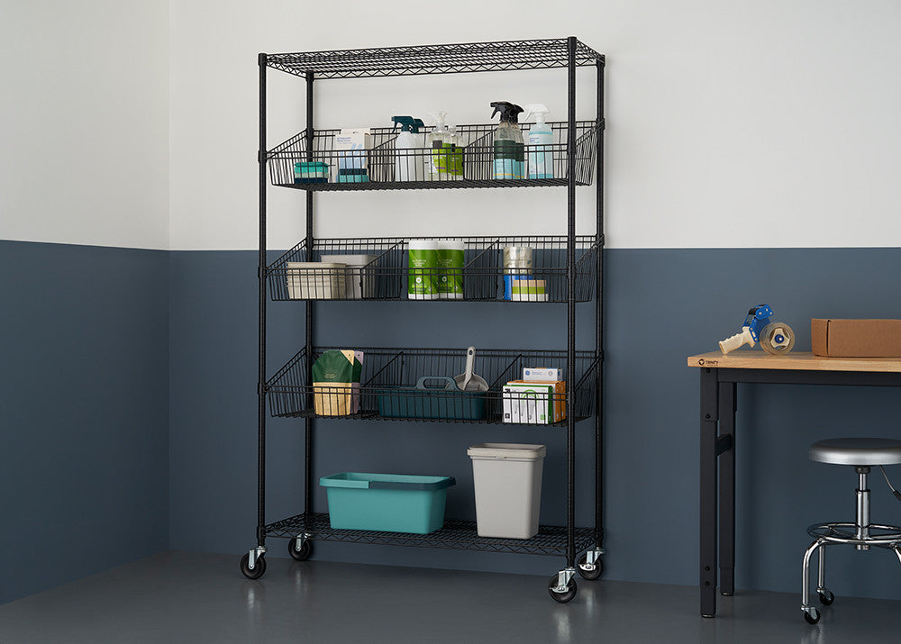 A black, five-tier wire shelving unit on wheels stands against a two-tone blue and white wall, holding various storage bins and items. A wooden table with a stool and packing supplies is visible to the right.