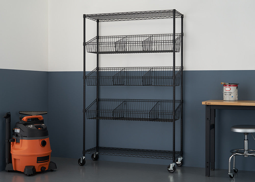 A black wire shelving unit with five tiers, each containing a wire basket and dividers, stands against a two-toned wall. The shelves are on wheels, and a wet/dry vacuum and a small table with a stool are visible in the background.
