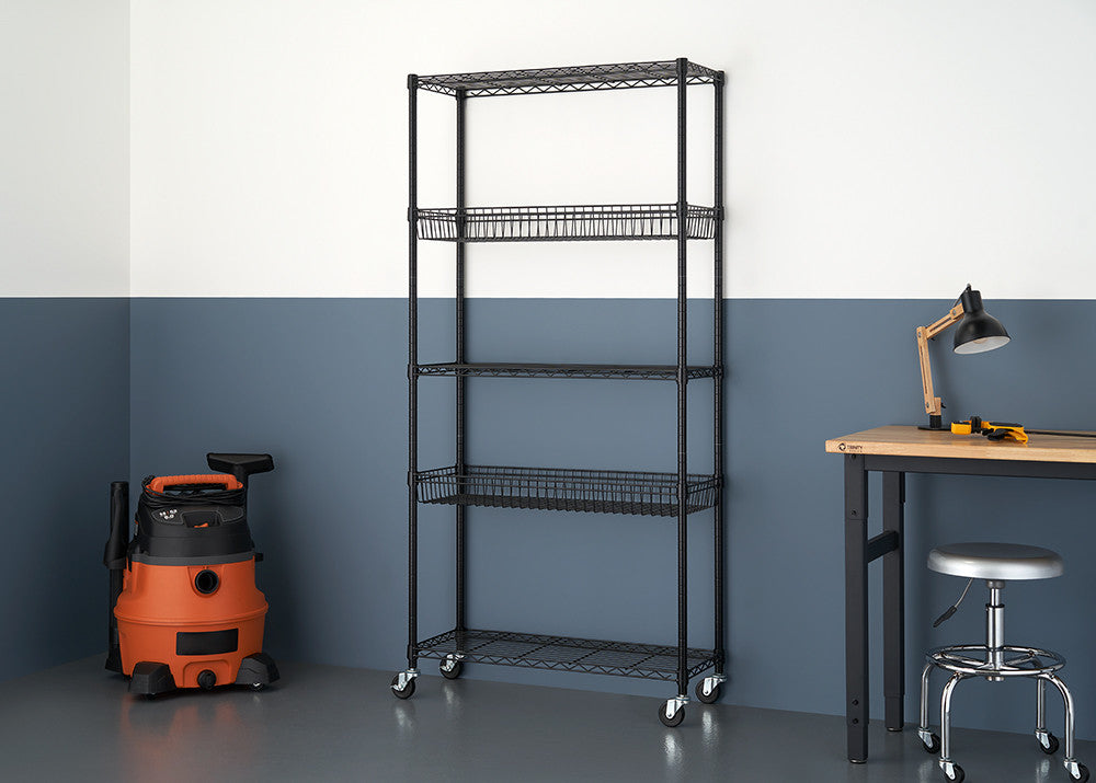 A black wire shelving unit with five tiers stands against a two-toned wall, featuring two wire basket shelves and three flat wire shelves. The unit is on casters and is positioned between an orange wet/dry vacuum and a wooden desk with a lamp and stool.
